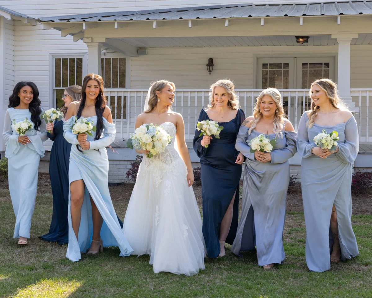 A group of women, including a bride in a white wedding gown, standing outdoors in front of a house, holding white bouquets, smiling and talking.