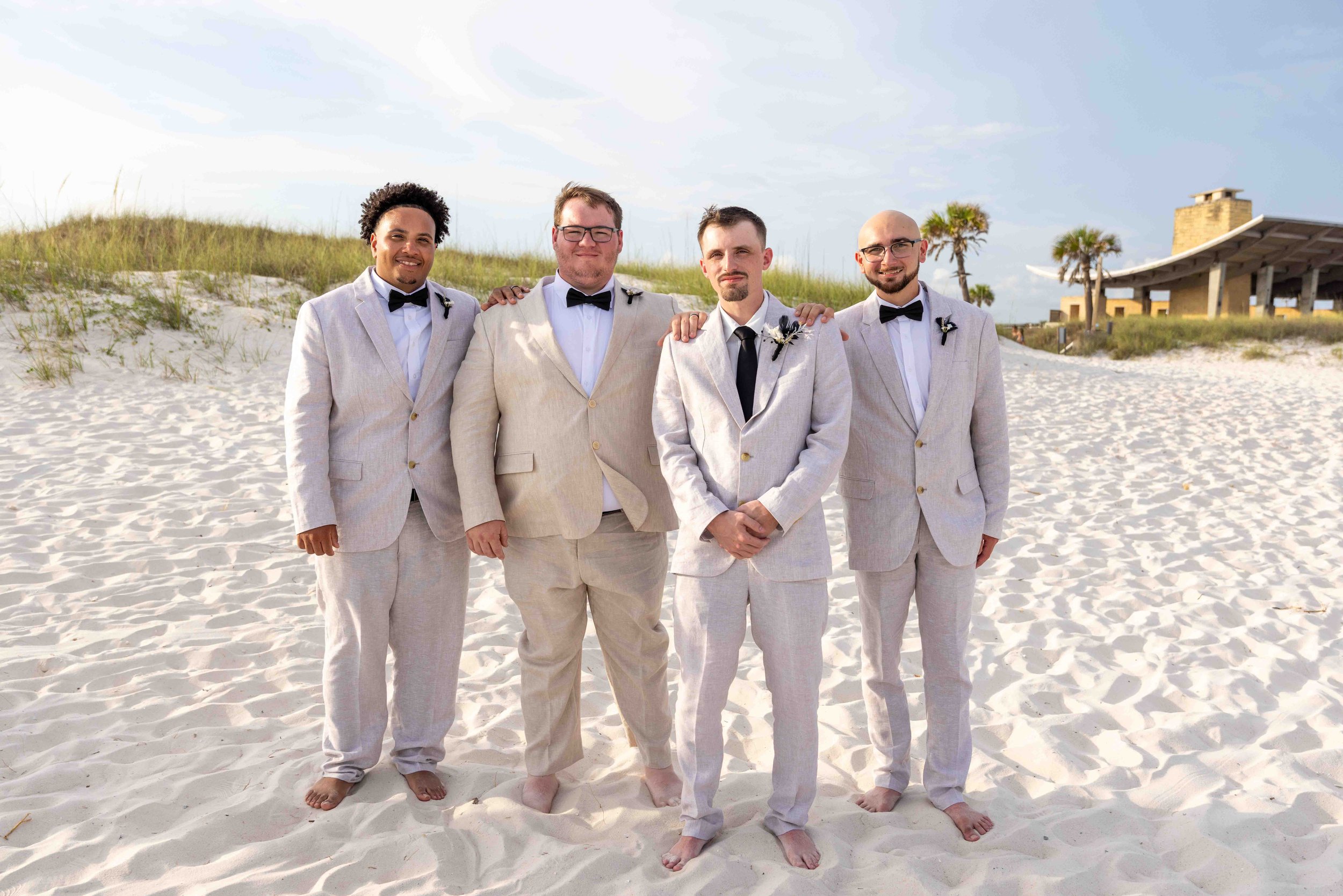 Group of four men in wedding suits standing on a sandy beach with grass dunes and a small pavilion in the background.