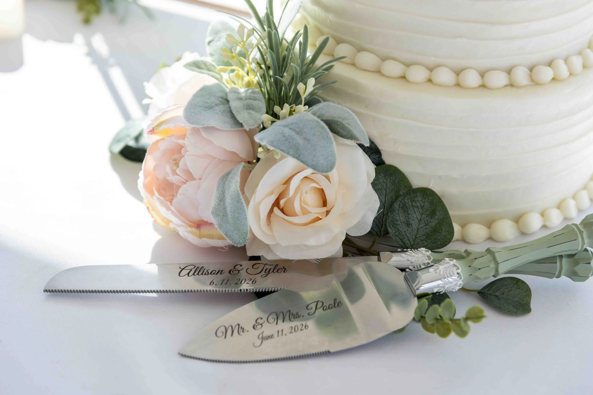 A wedding cake decorated with white frosting and a floral arrangement of pink and white roses, green leaves, and silver-gray foliage, with a tag reading 'Mr. & Mrs. Paige, June 11, 2026'.