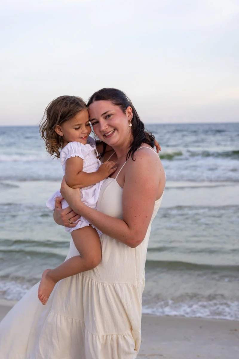 A woman and a young girl are smiling and hugging at the beach, with the ocean and sky in the background.