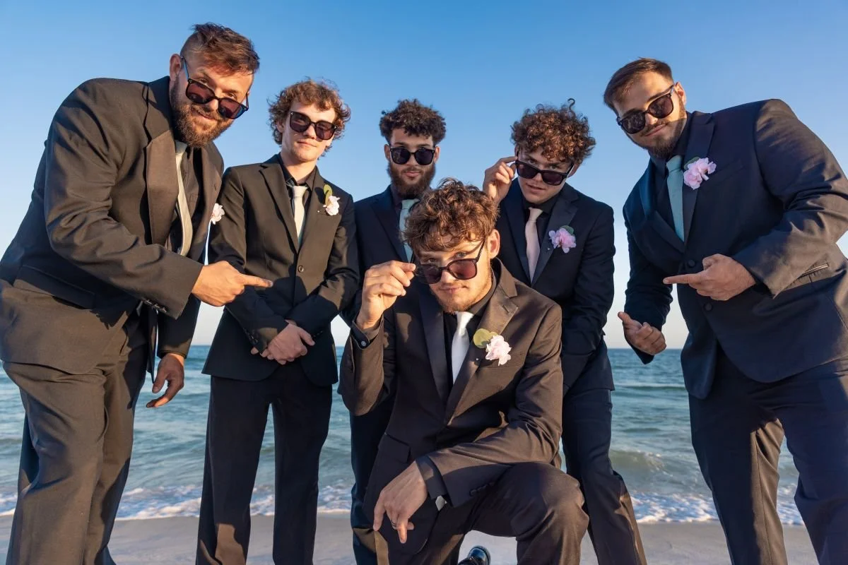 Group of six men in suits and sunglasses posing on the beach, some pointing and one squatting in the front.