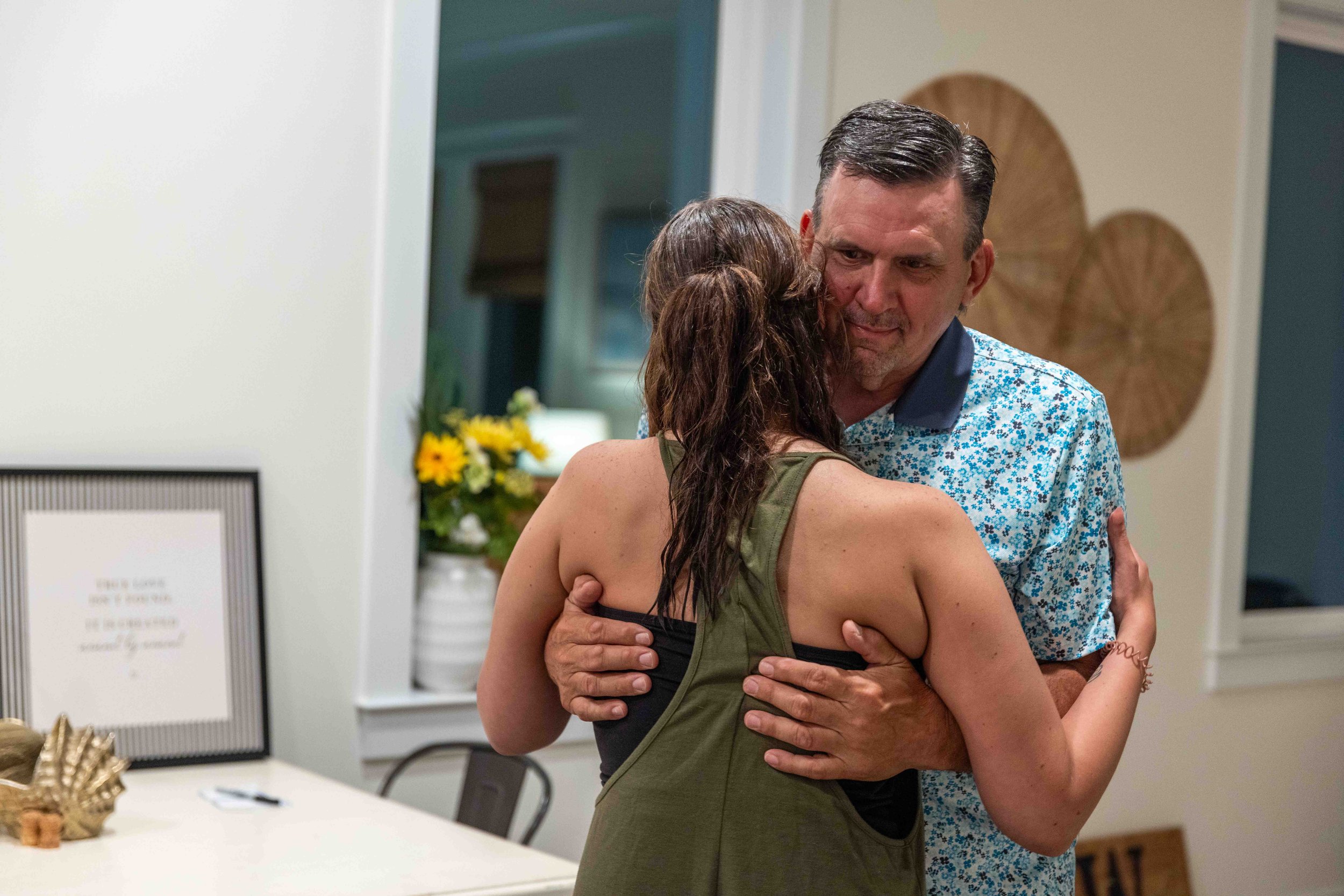 A man and a woman embracing in a hug indoors near a table with decor and yellow flowers.