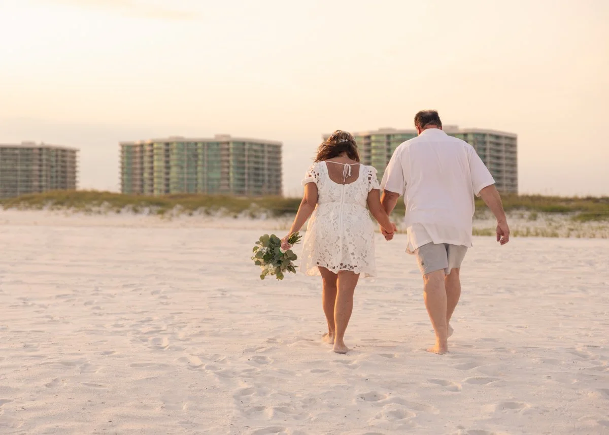 Couple walking hand in hand on a sandy beach during sunset, with buildings in the background. The woman wears a white lace dress and holds a bouquet of flowers, while the man wears a white shirt and shorts.