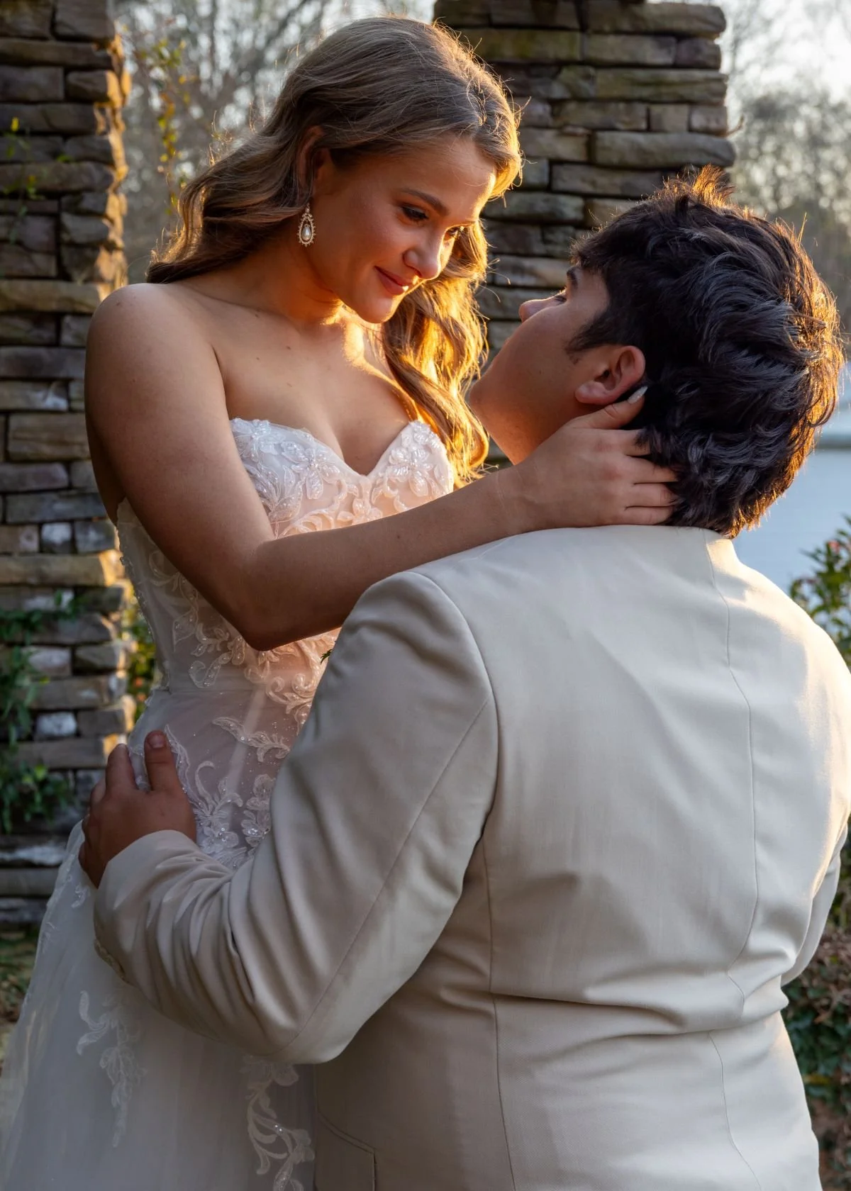 A bride and groom embrace outdoors during sunset, with the bride wearing a strapless lace wedding gown and pearl earrings, and the groom in a light-colored suit, near a stone wall with trees in the background.