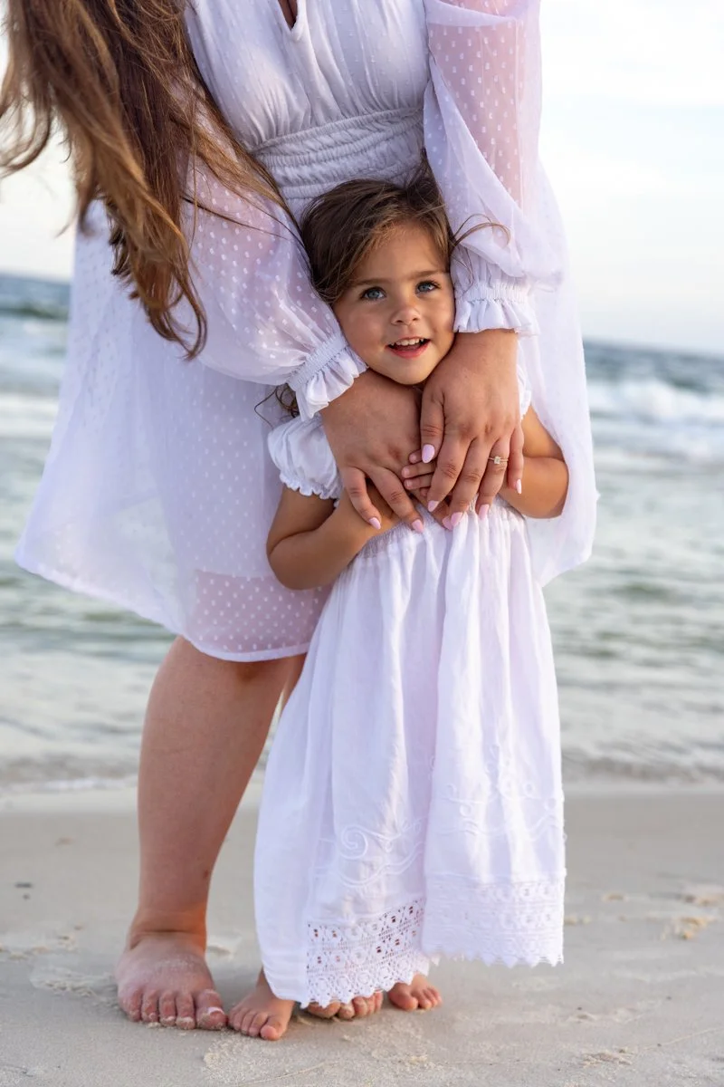 A woman and a young girl standing on the beach, with the woman embracing the girl from behind. Both are wearing white dresses. The ocean waves are visible in the background.