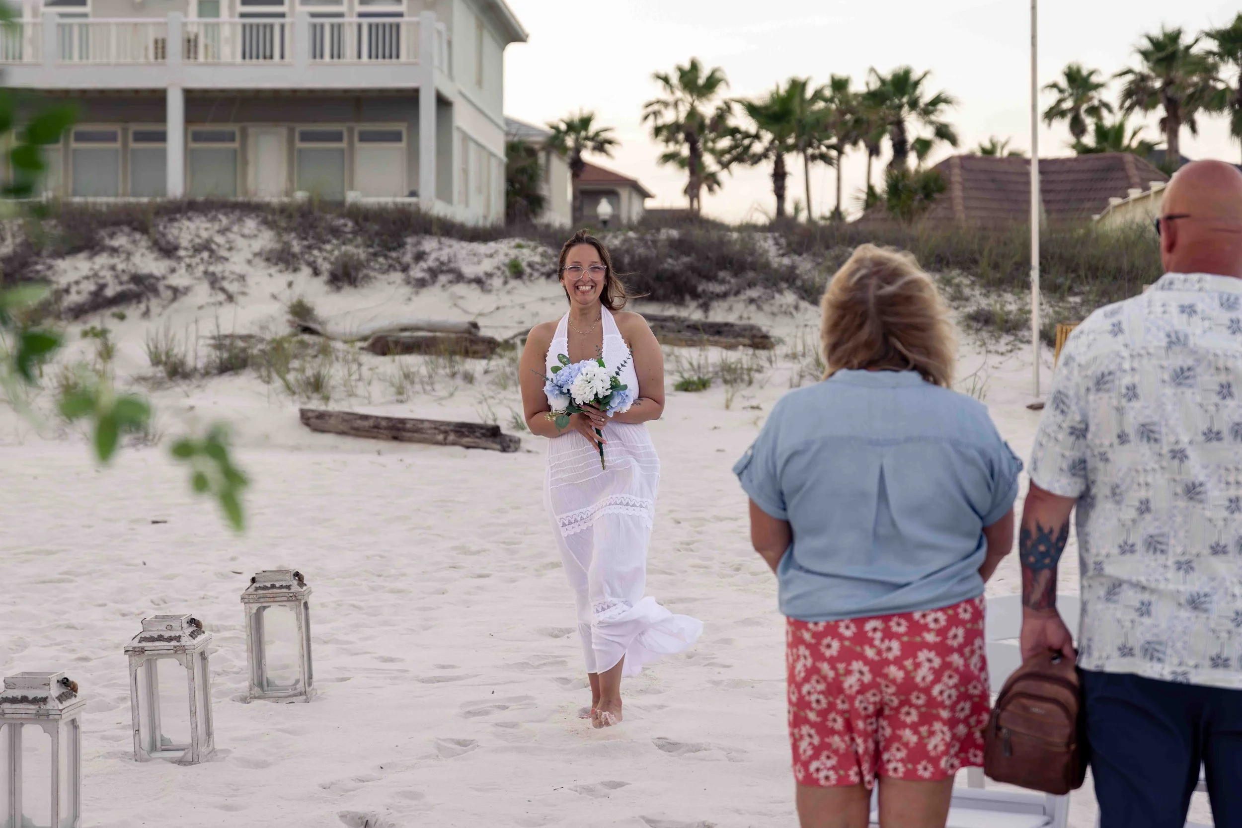 A woman in a white dress holding a bouquet of flowers, smiling, standing barefoot on a sandy beach during a small outdoor wedding ceremony. There are three people facing away in the foreground, and houses with palm trees are in the background.