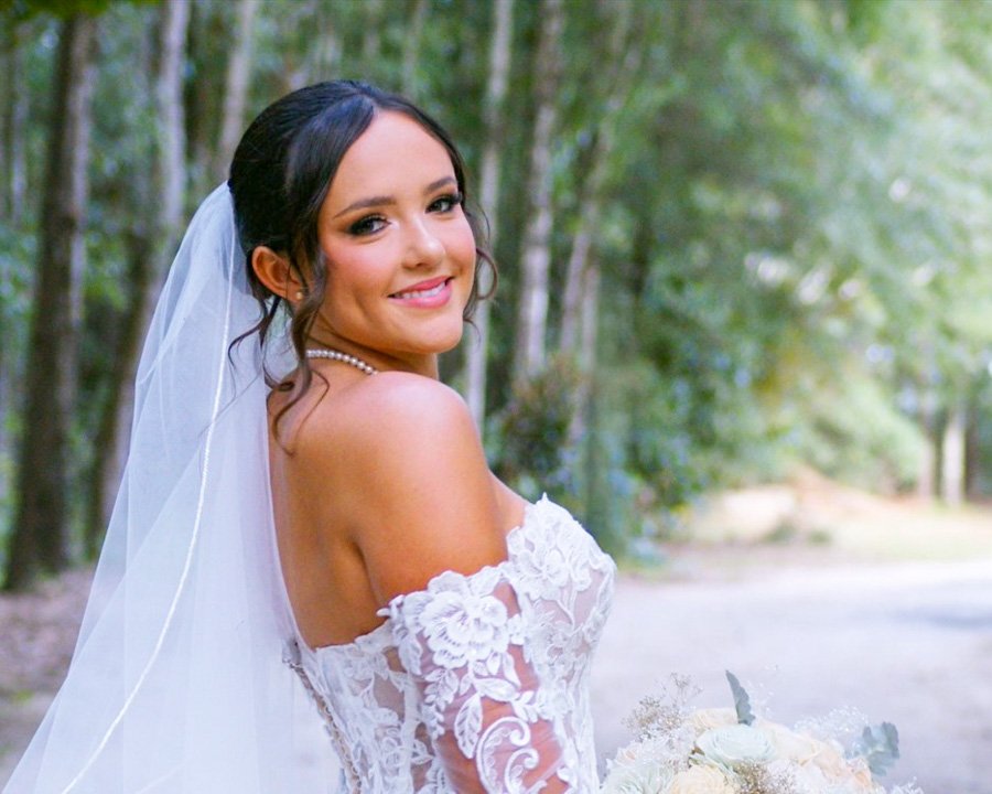 A smiling bride with dark hair in an updo, wearing a strapless white lace wedding dress and a pearl necklace, holding a bouquet of white flowers, outdoors with green trees in the background.