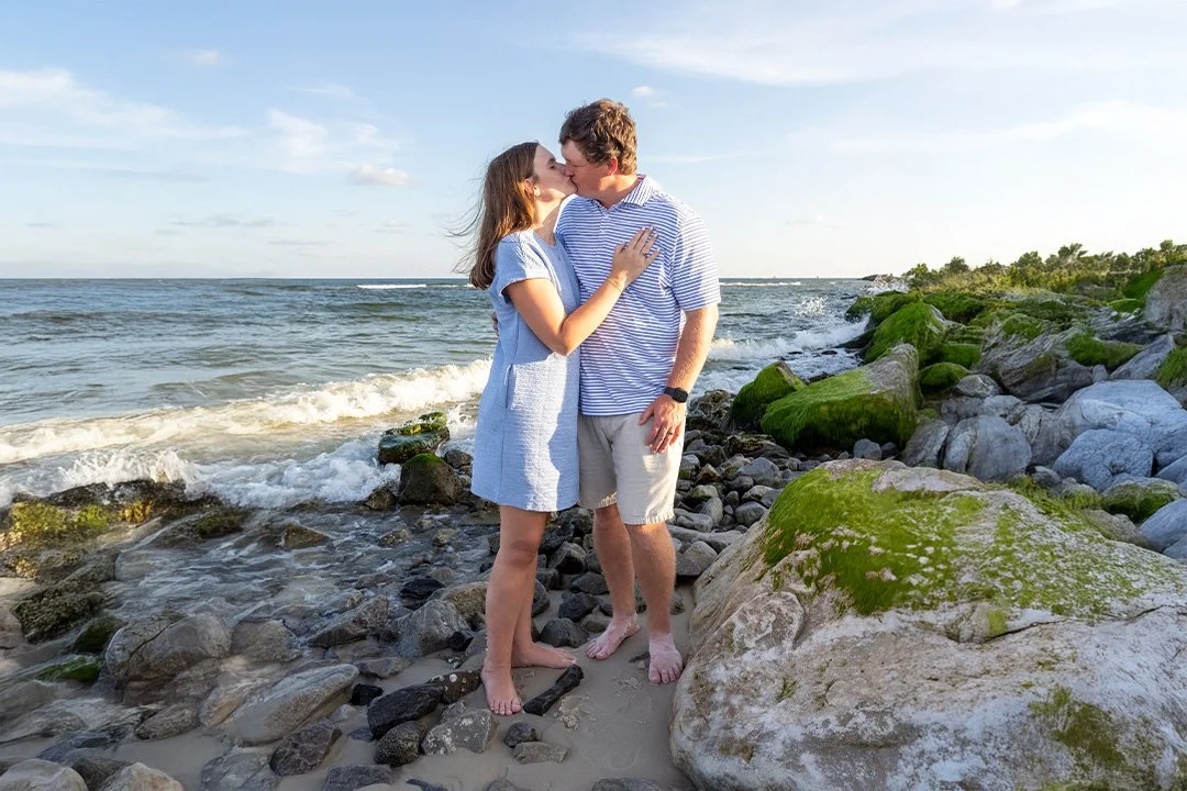A young couple sharing a kiss on a rocky beach with waves and moss-covered rocks in the background.