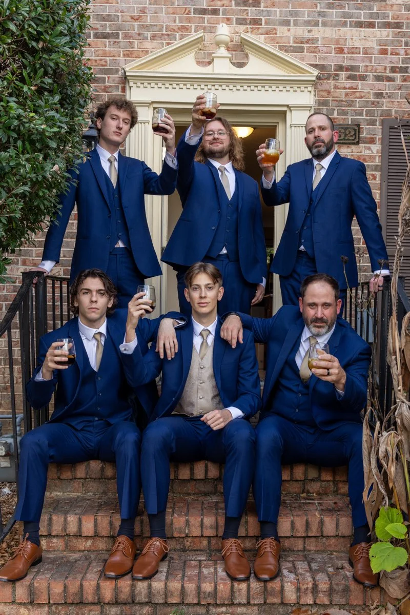 Six men dressed in blue suits and cream waistcoats, holding glasses of dark-colored drinks, standing and sitting on a brick staircase in front of a brick house, raising glasses in a toast