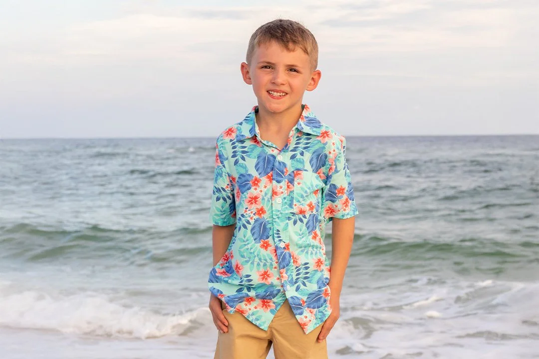 A young boy standing on the beach with ocean waves behind him, wearing a colorful floral short-sleeve shirt and beige shorts.