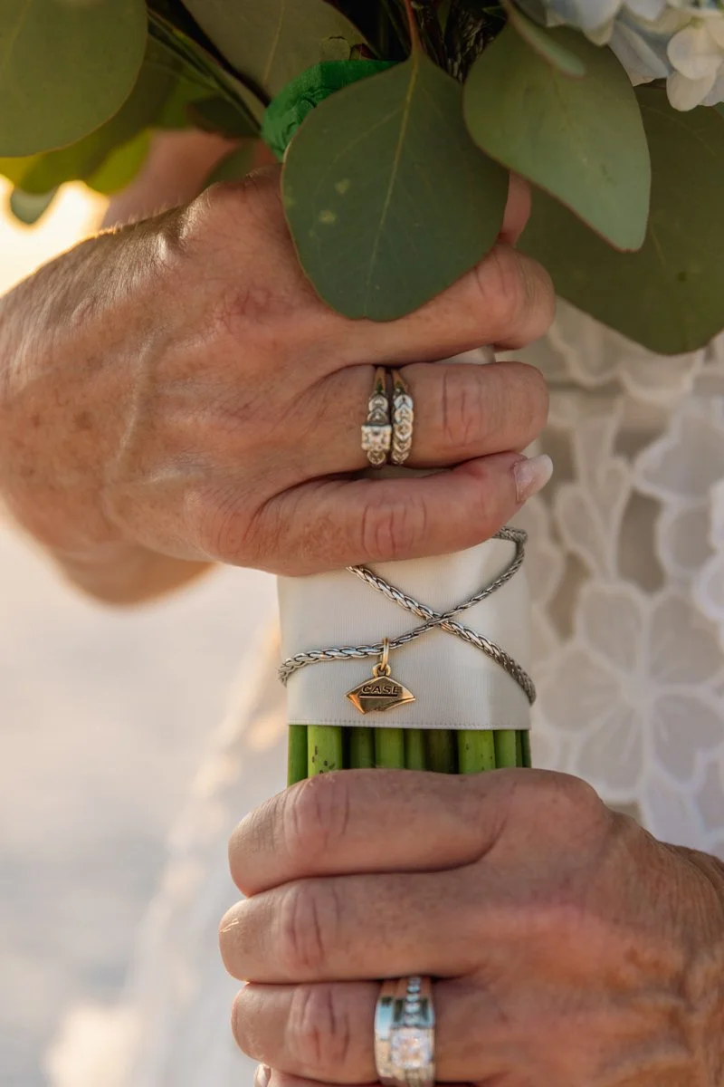 Close-up of elderly hands holding a bouquet of flowers wrapped with a white ribbon, adorned with rings and bracelets.