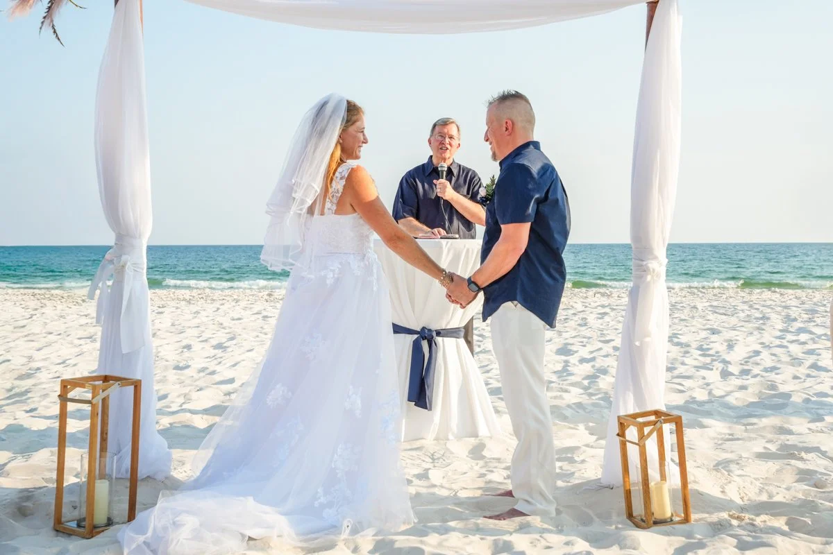 A couple gets married on the beach during the daytime. They are holding hands and looking at each other as they exchange vows, under a white canopy. An officiant stands behind a small table, speaking into a microphone. The scene is set against the oc