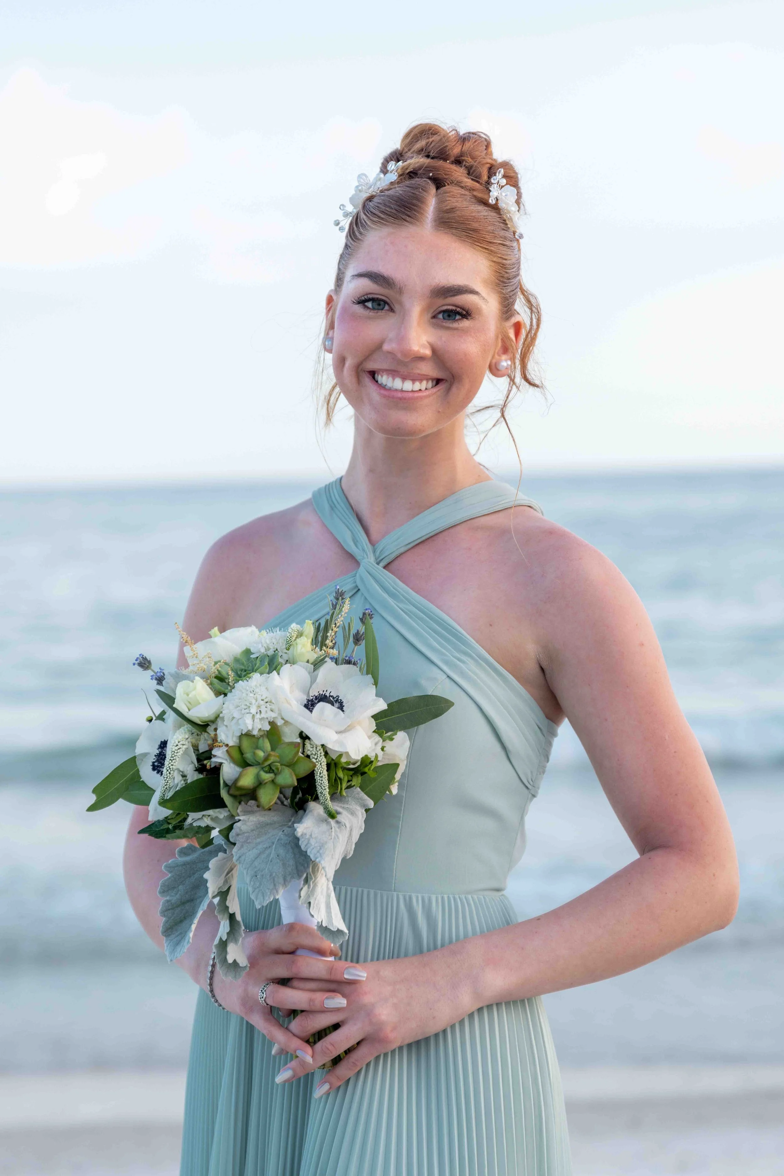 A smiling woman with red hair styled in an updo, wearing a light blue dress with a crisscross neckline, standing on a beach with the ocean and sky in the background, holding a bouquet of white flowers and greenery.