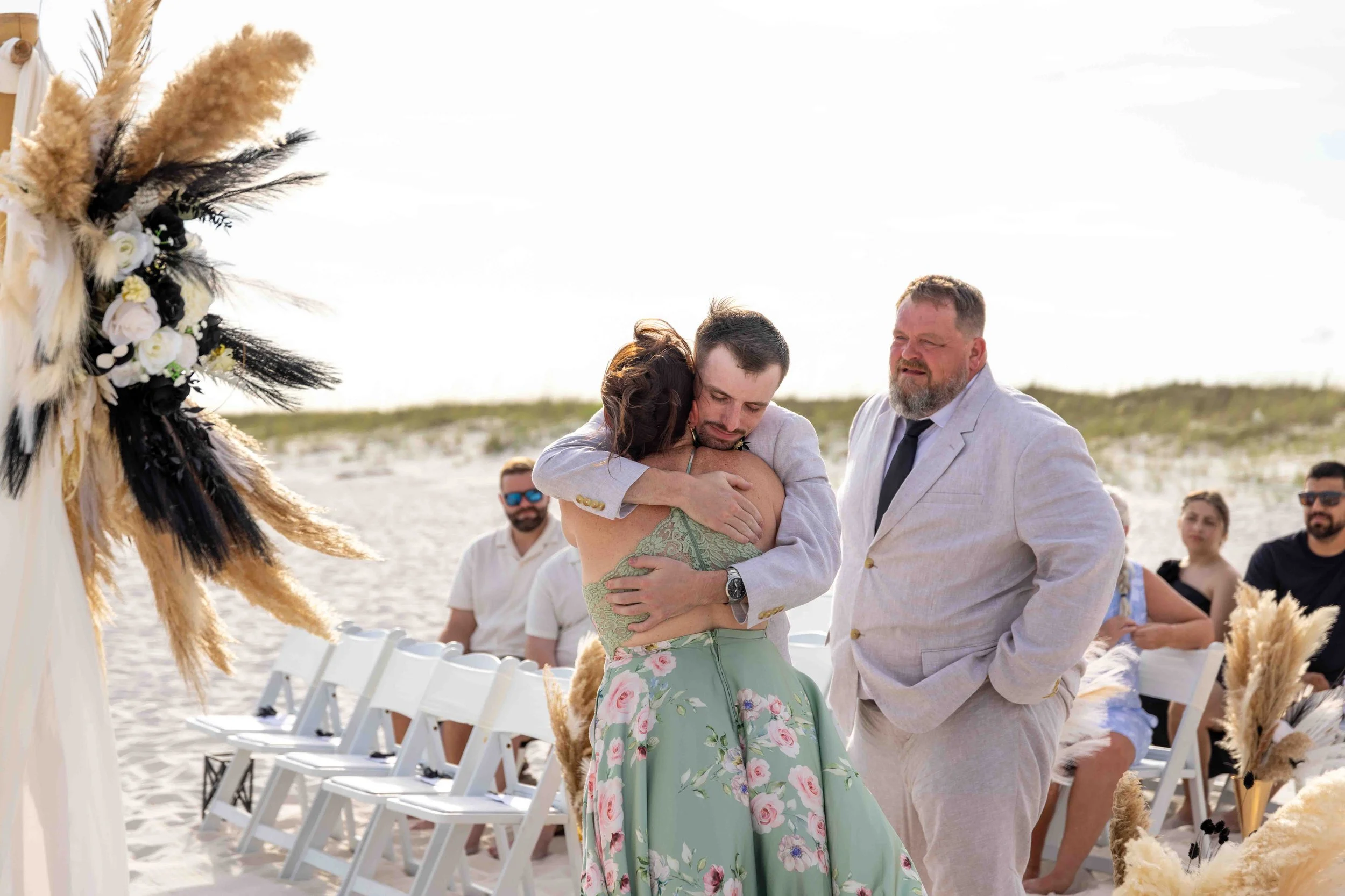 A beach wedding ceremony with friends and family, showing a man in light-colored suit embracing a woman in a green floral dress, and another man in a light-colored suit smiling nearby, with white folding chairs and decorative feathers and flowers.
