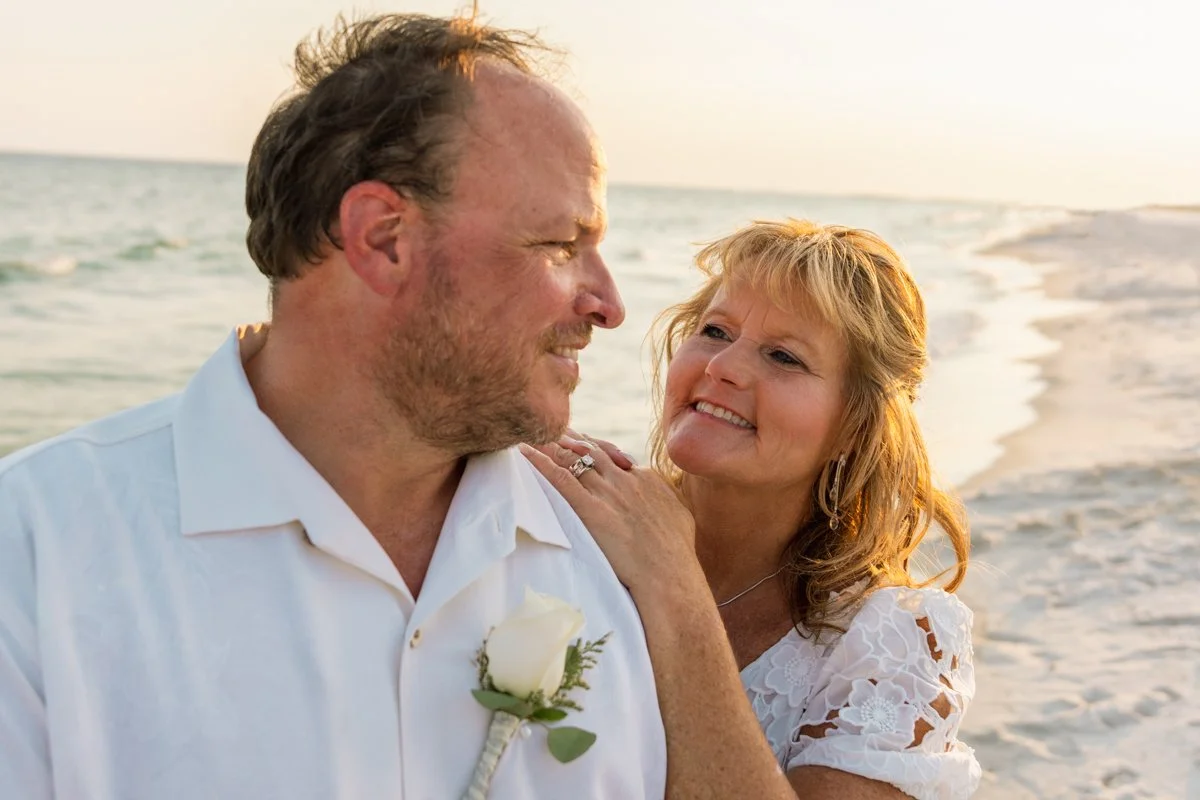 A middle-aged couple at the beach, dressed in white, smiling and enjoying a tender moment during sunset.
