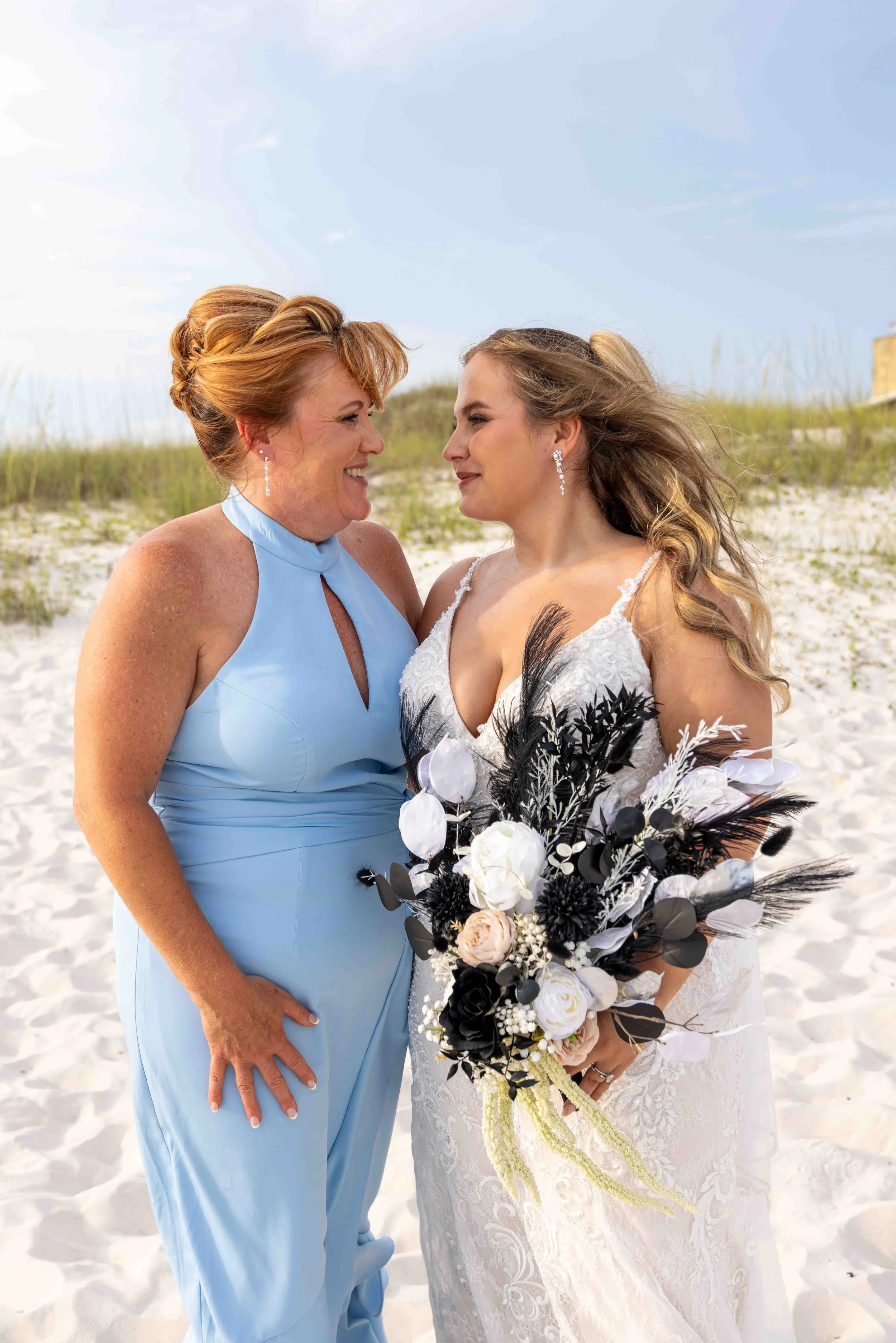 Two women, one in a light blue dress and the other in a white wedding gown, smile at each other on a sandy beach with green dunes and a blue sky in the background. The bride holds a bouquet of white and black flowers with black, white, and pink accen