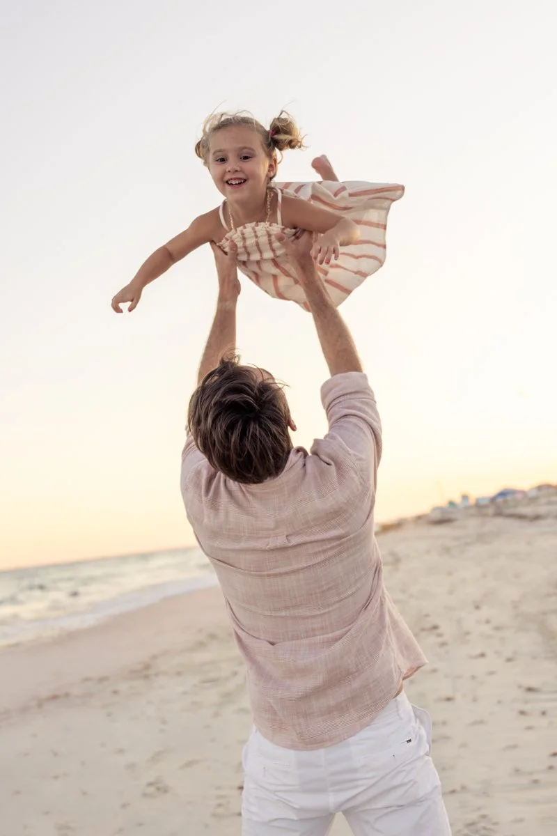 A man is lifting a young girl in the air on the beach during sunset, both smiling and enjoying the moment.