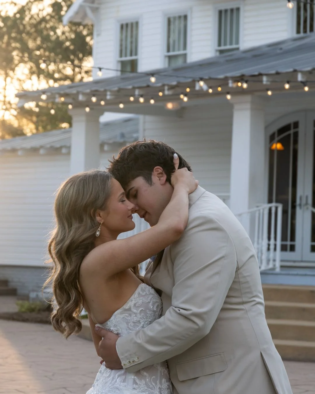 A romantic moment between a bride and groom during their wedding, embracing in front of a white house decorated with string lights at sunset.
