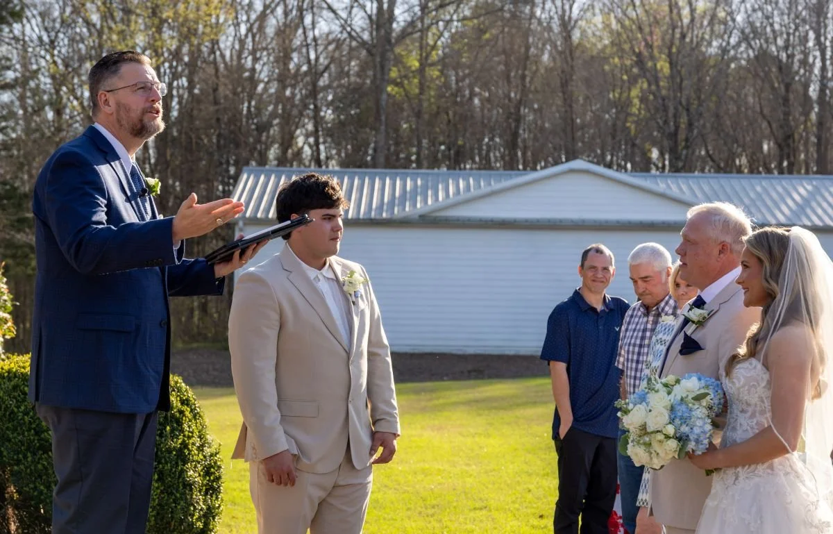 A wedding ceremony outdoors with a bride in a white wedding dress holding a bouquet, an elderly groom, a young man in a cream suit, and a man in a blue suit giving a speech, with three men in casual shirts watching.