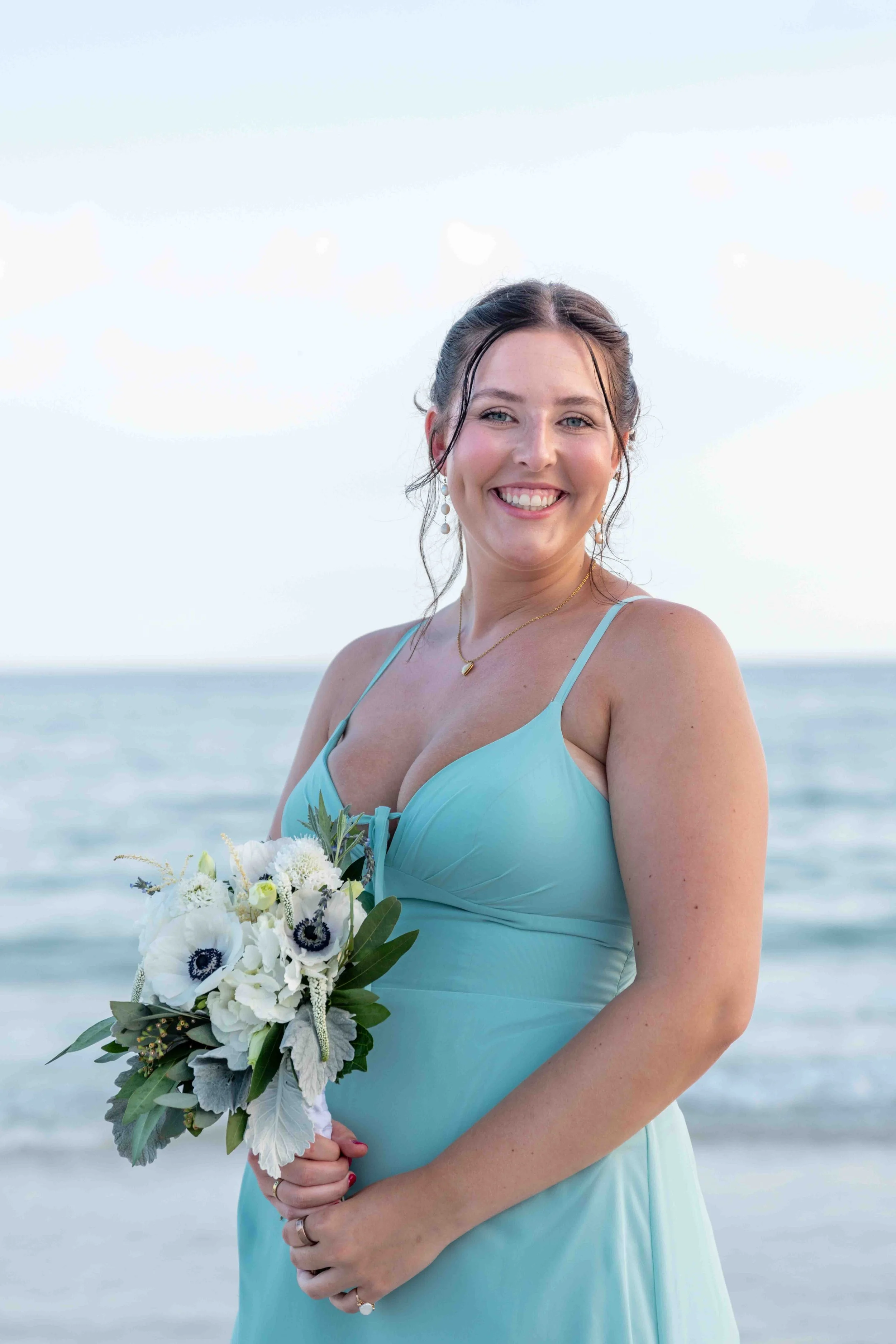 A smiling woman in a light blue dress holding a bouquet of white and purple flowers at the beach.