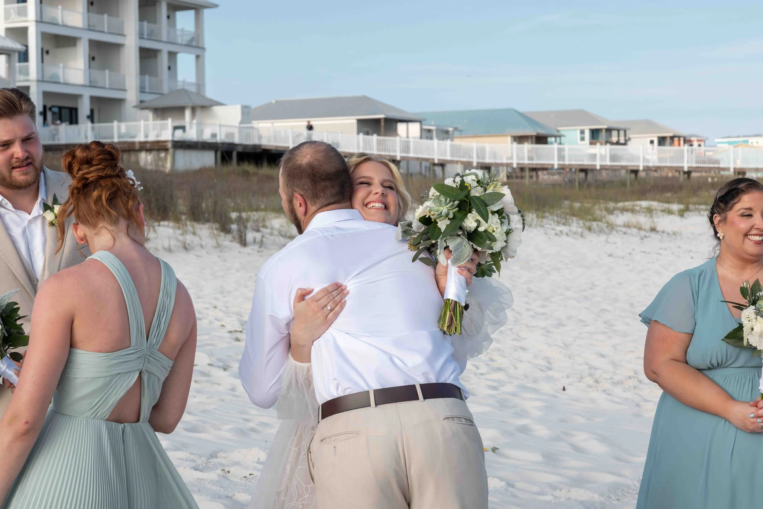 A group of people celebrating on a beach, with a man in beige pants and a woman holding a bouquet hugging, and others smiling nearby. There are beach houses in the background.