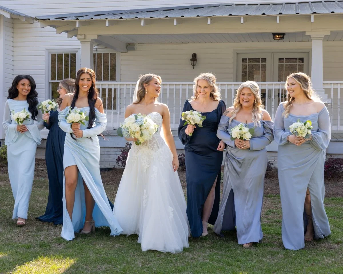 A group of women, including a bride in a white wedding gown, and bridesmaids in various shades of blue dresses, standing outside a white house with a porch, holding bouquets of white flowers, smiling and walking together.