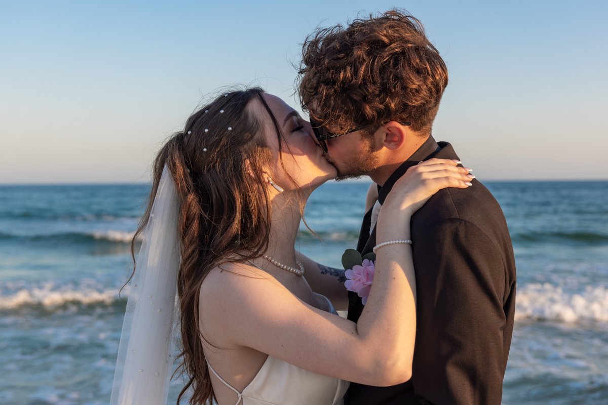 A bride and groom kissing on the beach during sunset, with the ocean and sky in the background.