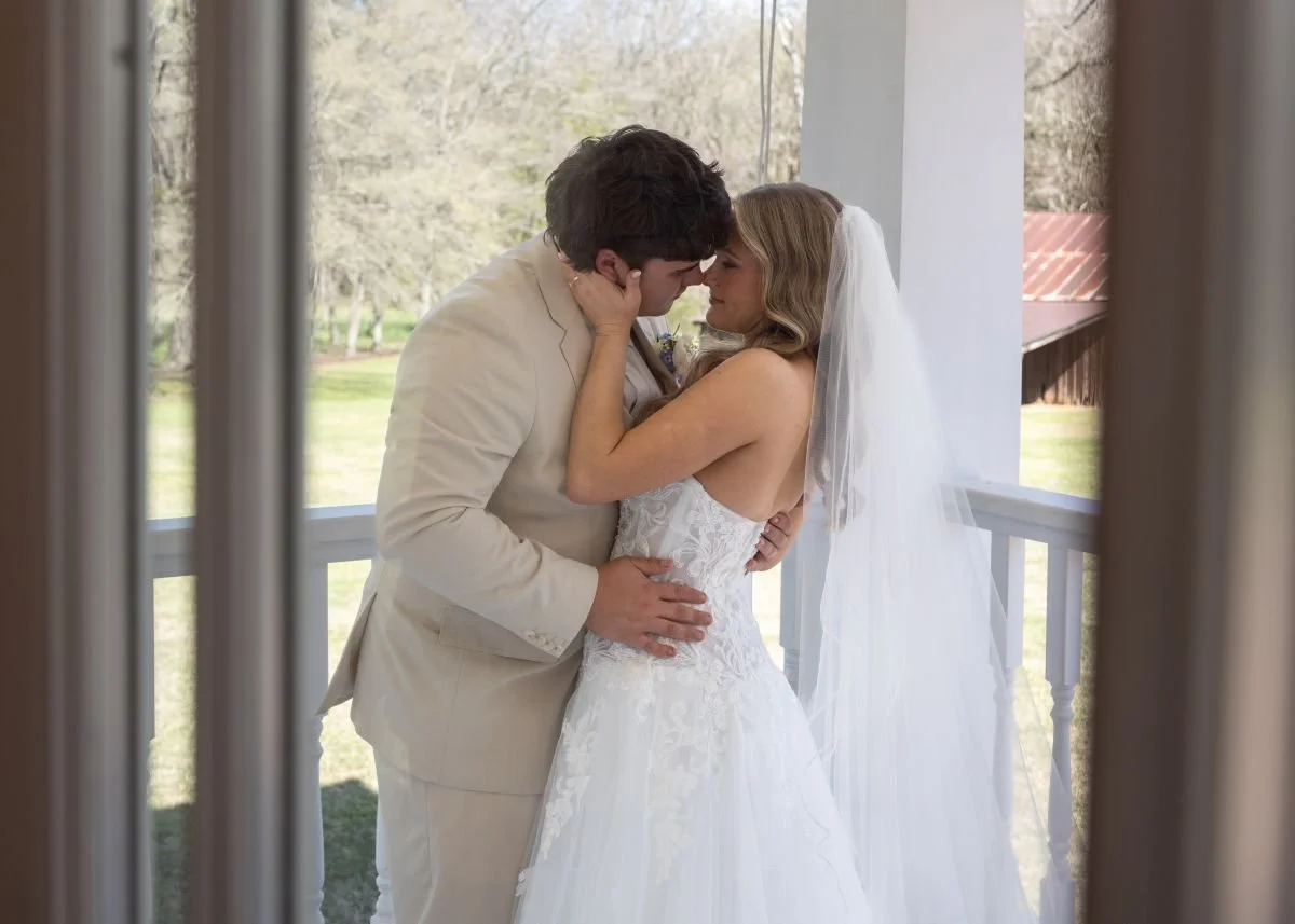 A newlywed couple sharing an intimate moment indoors, with the bride in a strapless lace wedding gown and veil, and the groom in a cream-colored suit, standing close together as they touch foreheads.