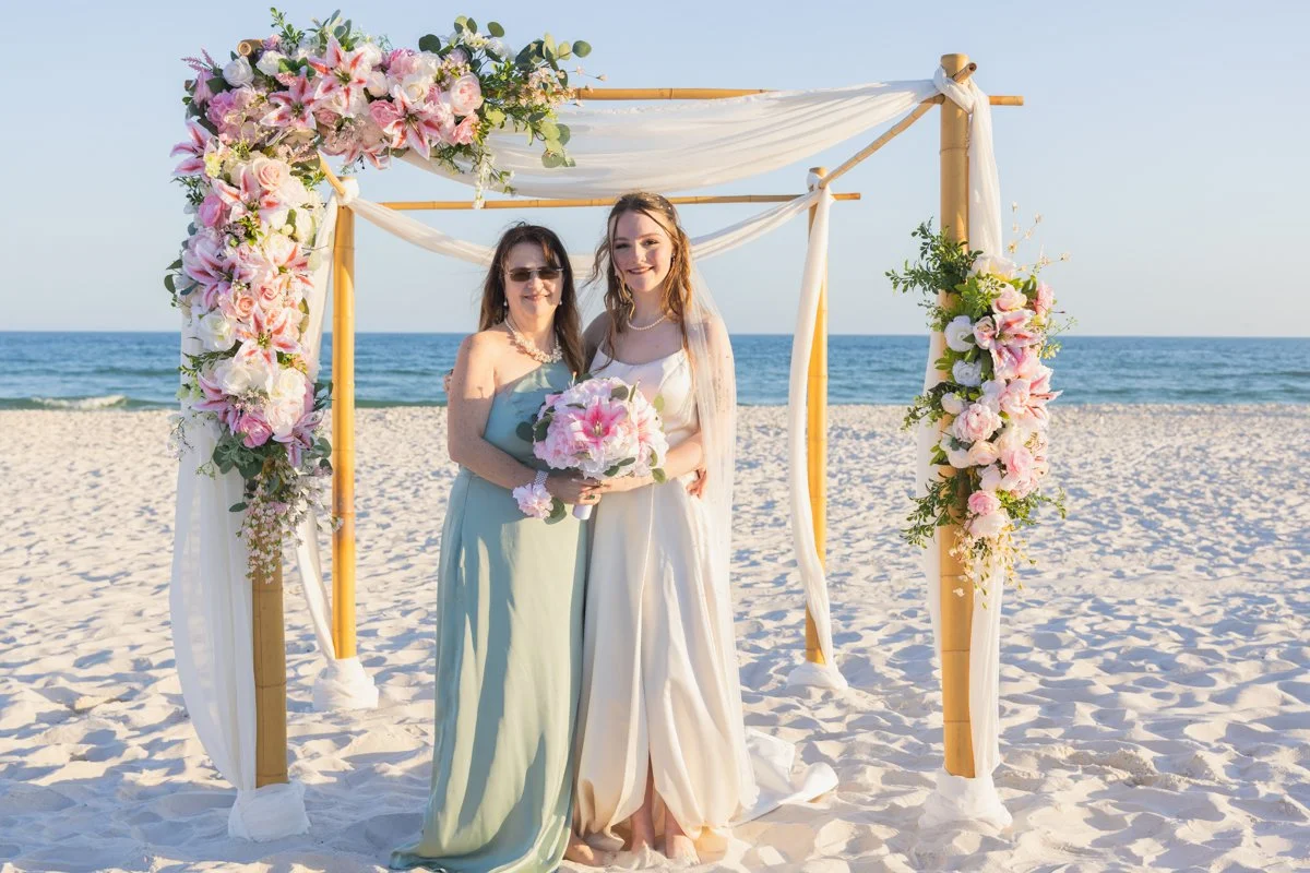 Two women standing under a wedding arch decorated with pink and white flowers on a beach, with the ocean and sky in the background.