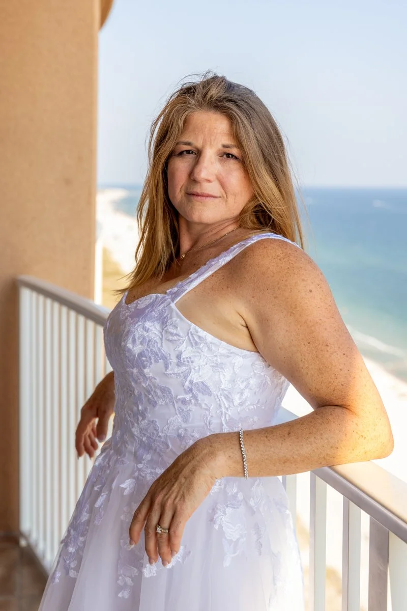A middle-aged woman with long, light brown hair stands on a balcony by the beach, wearing a white dress with floral embroidery and looking at the camera.
