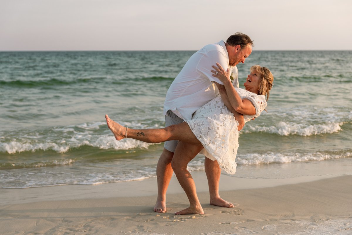 A couple dancing and embracing on a beach near the ocean at sunset, with the man lifting the woman who is smiling and wearing a white lace dress.