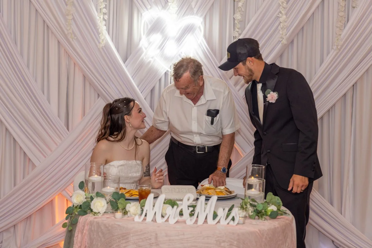 A bride and groom smiling at an elderly man during their wedding reception. The couple is sitting at a decorated table with white flowers, candles, and a 'Mr & Mrs' sign. The background features draped white fabric with hearts and soft lighting.