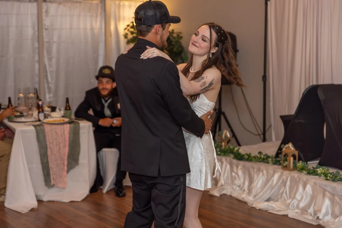 A couple is dancing at their wedding reception, with the bride in a white dress and the groom in a black suit. They are holding each other and smiling, with guests seated at tables in the background.