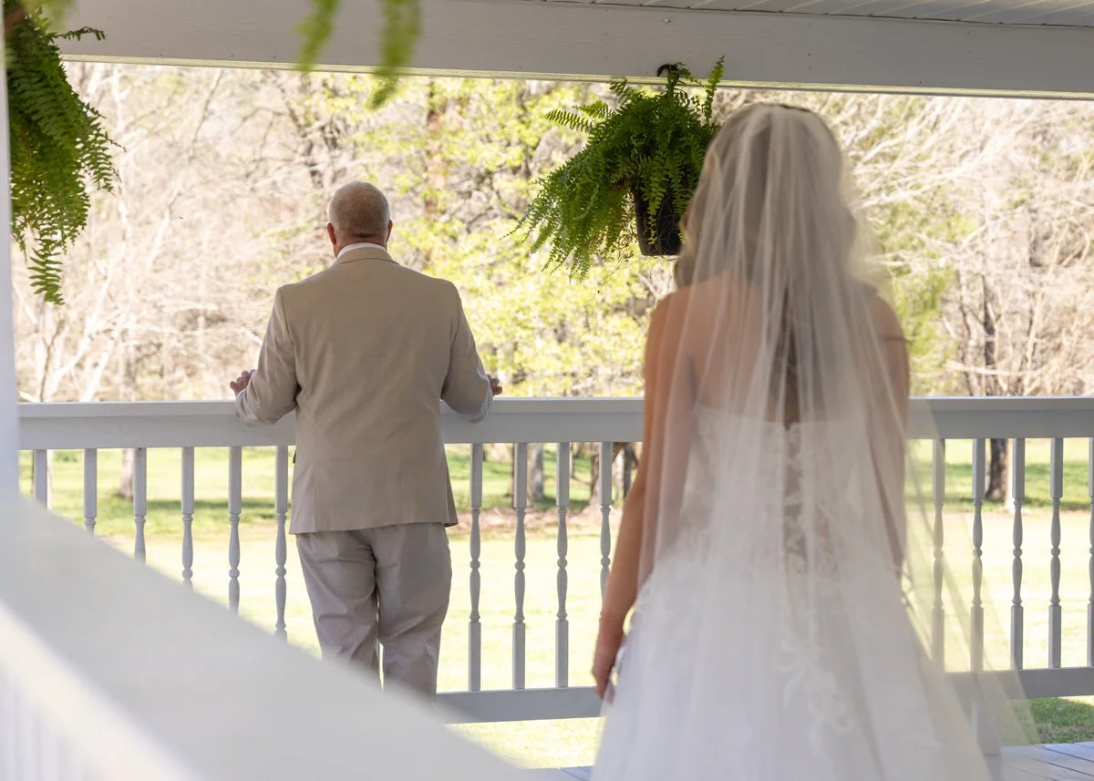 A bride wearing a wedding dress with a veil stands on a porch, watching a man in a light-colored suit who is standing and looking outside at a scenic outdoor area with trees and grass.