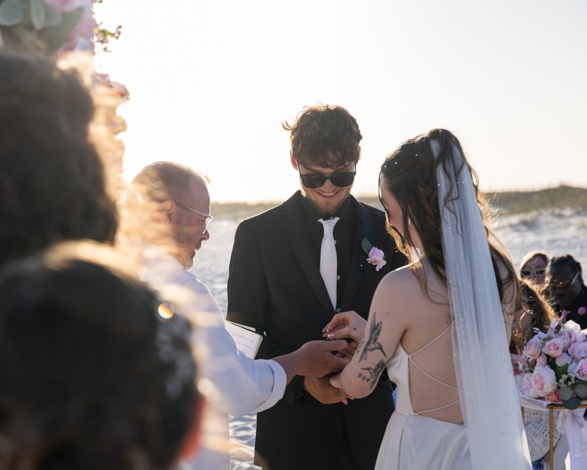 A wedding ceremony taking place on a beach during sunset, with a couple exchanging rings while holding hands, surrounded by officiants and guests.
