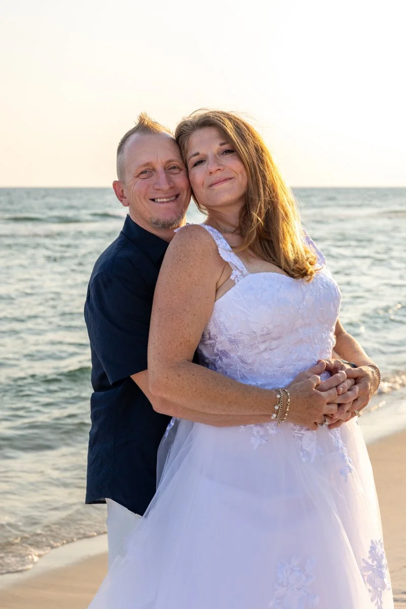 A couple embraces on the beach at sunset, with the ocean in the background. The woman wears a white dress, and the man wears a dark shirt.