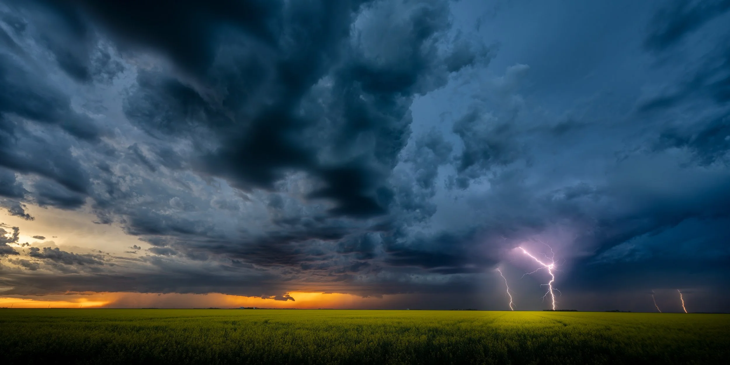 Dark storm clouds in the sky over a green field with lightning strikes visible near the horizon at sunset.