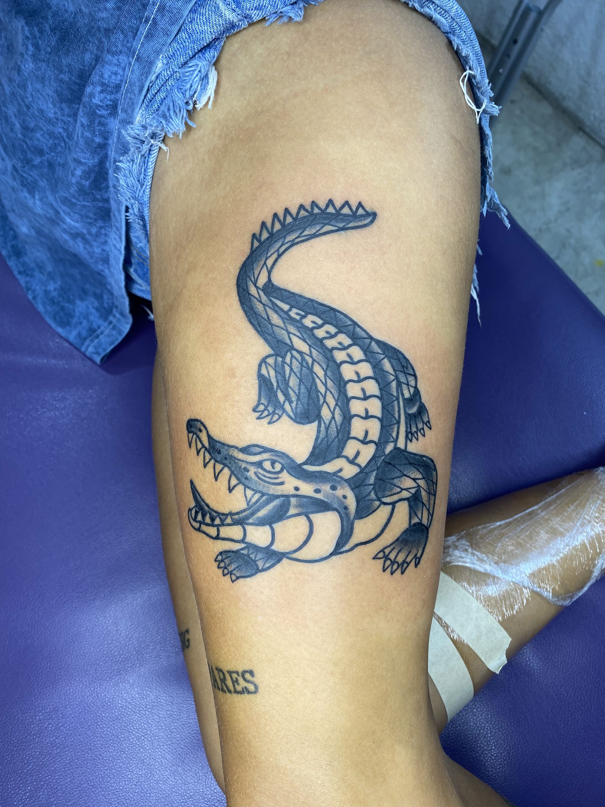 A person with a tattoo of a crocodile or alligator on their thigh, wearing ripped denim shorts.