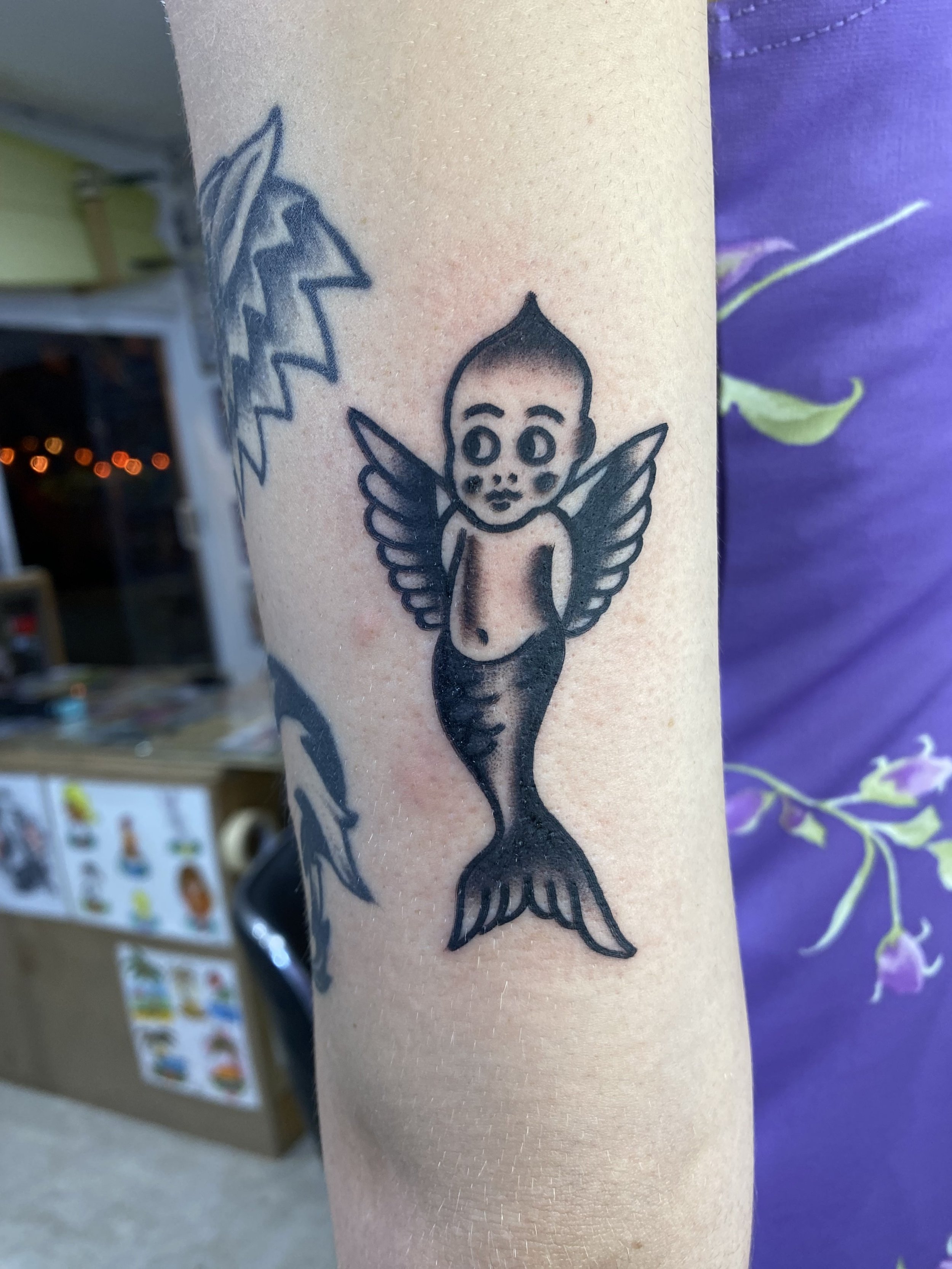 Tattoo of a cartoon character with big eyes and wings, holding a pitchfork, on a person's forearm.