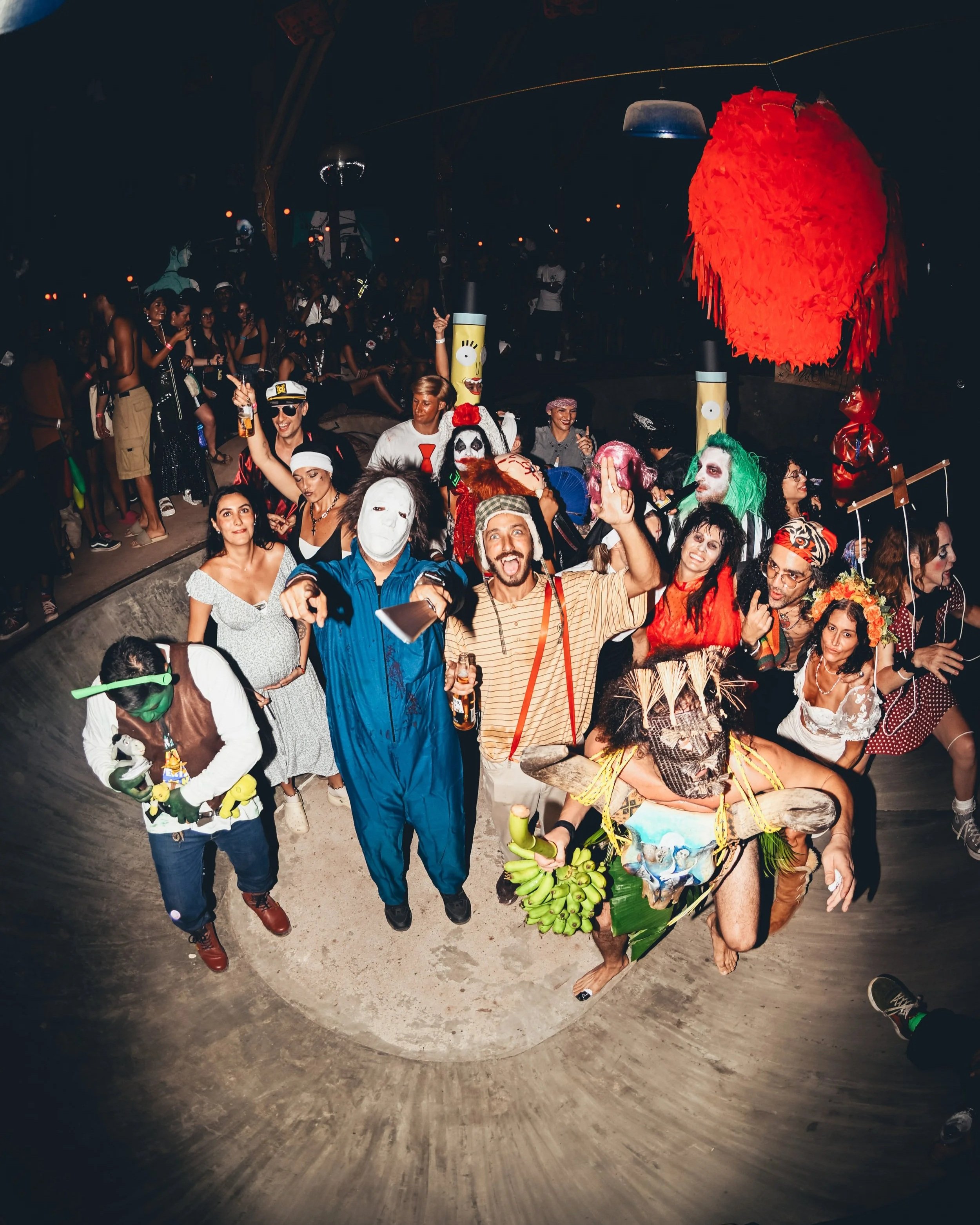 A large group of people at a party or celebration, many in costumes, with some wearing masks and face paint. Some are holding drinks and wearing accessories, with a big red paper lantern hanging above. The setting appears to be indoors with dim lighting.