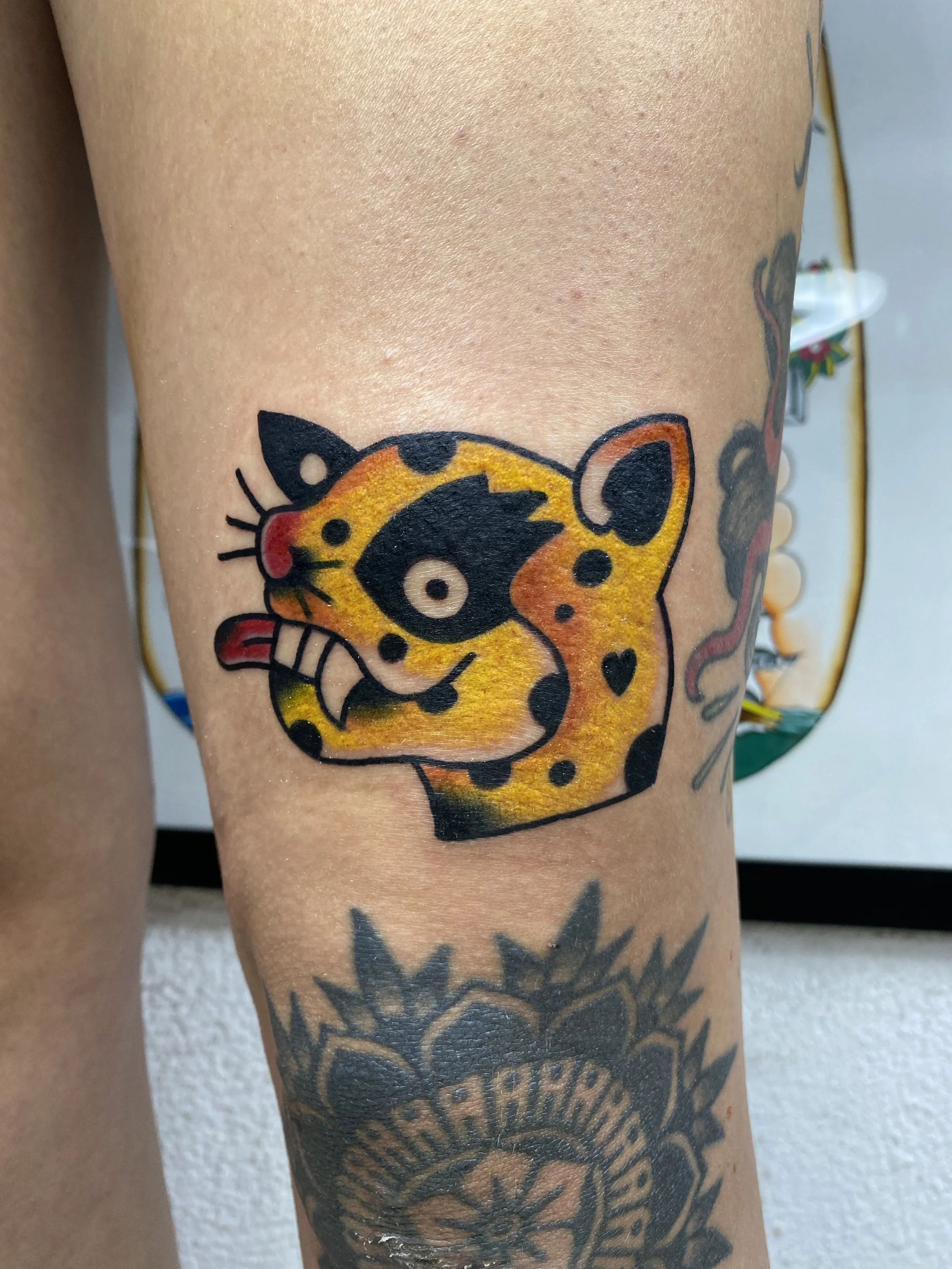 Close-up of a person's arm with a colorful tattoo of a stylized animal face, possibly a tiger or leopard, with bold black outlines and shades of yellow, orange, and red. The person's skin has a few small red spots and a larger dark tattoo beneath.