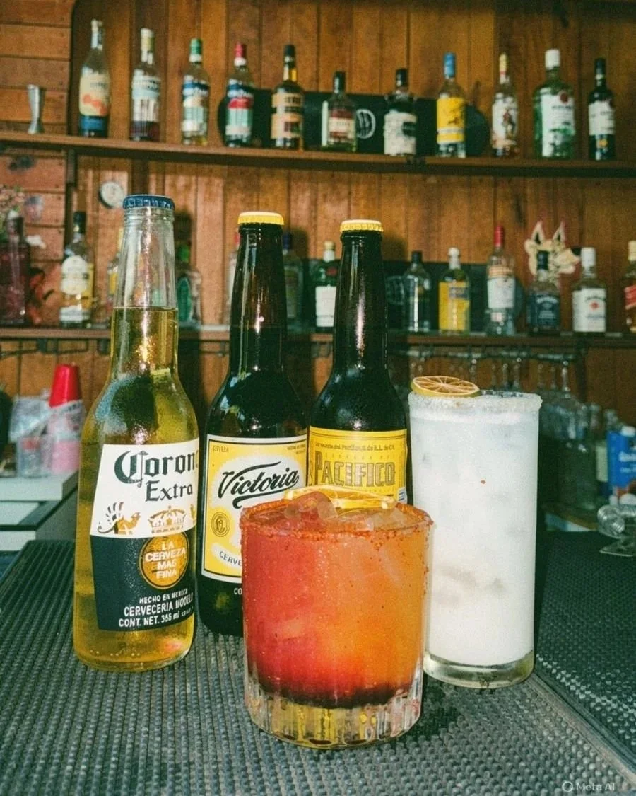 Three drinks on a bar counter: a Corona beer, two bottles of Victoria and Pacifico beers, a red cocktail with ice and lemon slice, and a tall white cocktail with a lemon garnish. Shelves with various liquor bottles in the background.