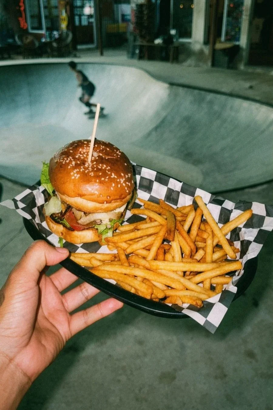 Person holding a basket of French fries and a cheeseburger with a skateboarder in a skate bowl in the background at night.