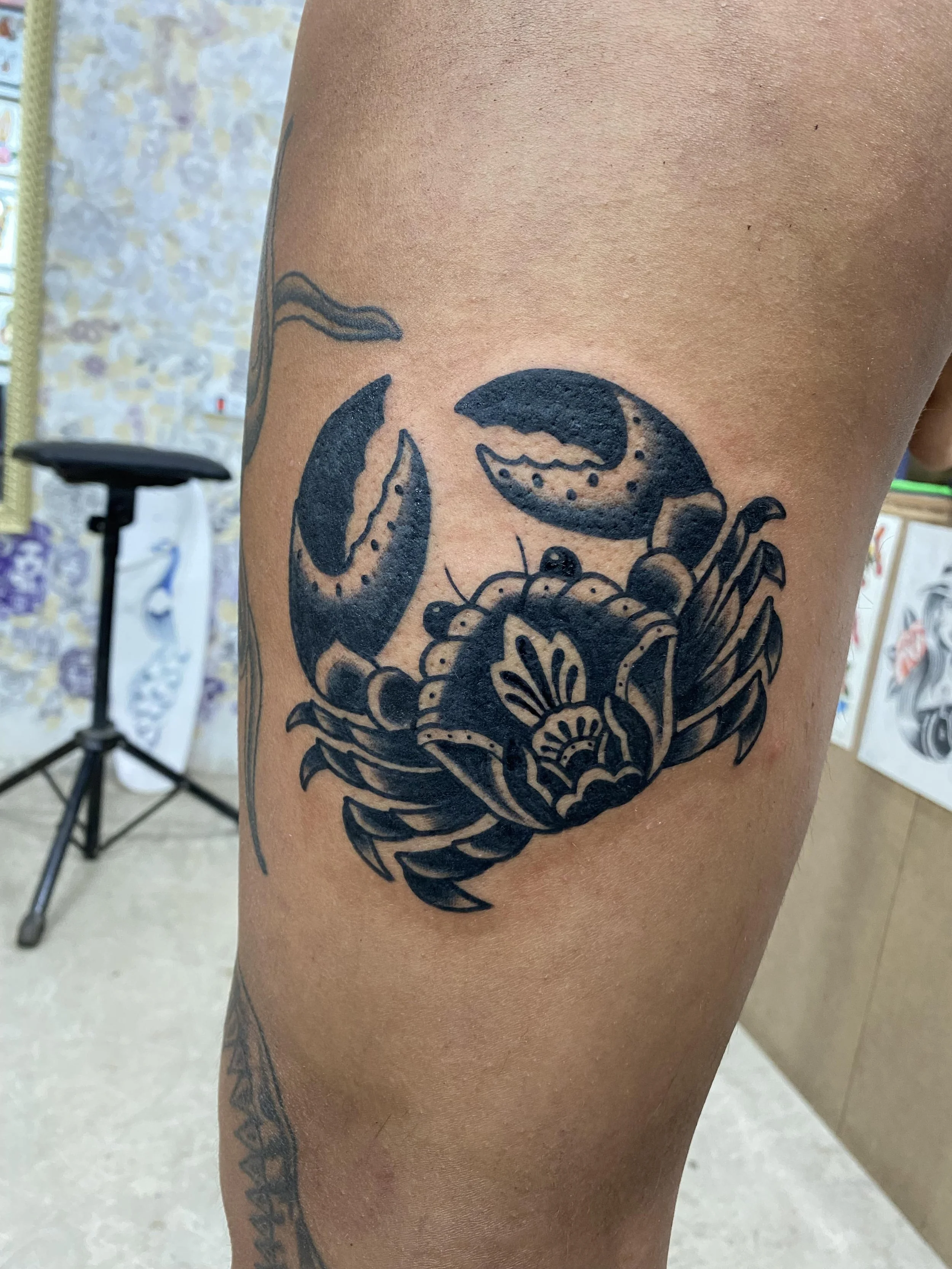 A tattoo of a crab with detailed claws and legs on a person's upper arm, with a blurred background showing a room with wall-patterned wallpaper and a stool.