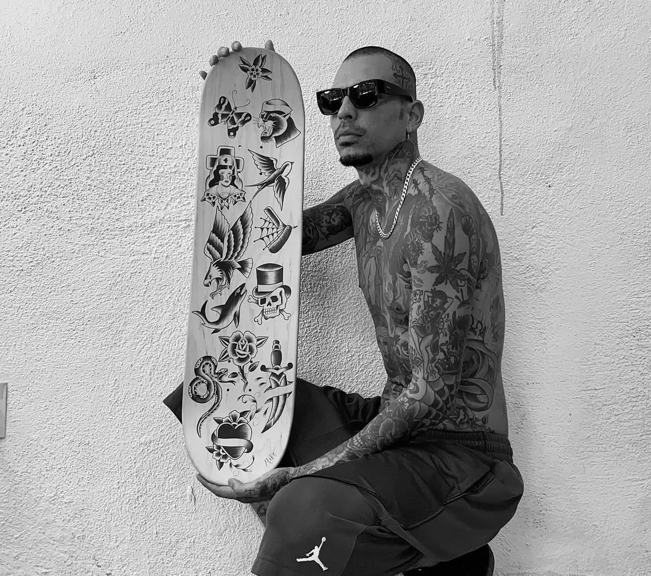 A tattooed man with sunglasses, wearing a chain, holding a skateboard with various tattoo-style images, kneeling against a textured wall.