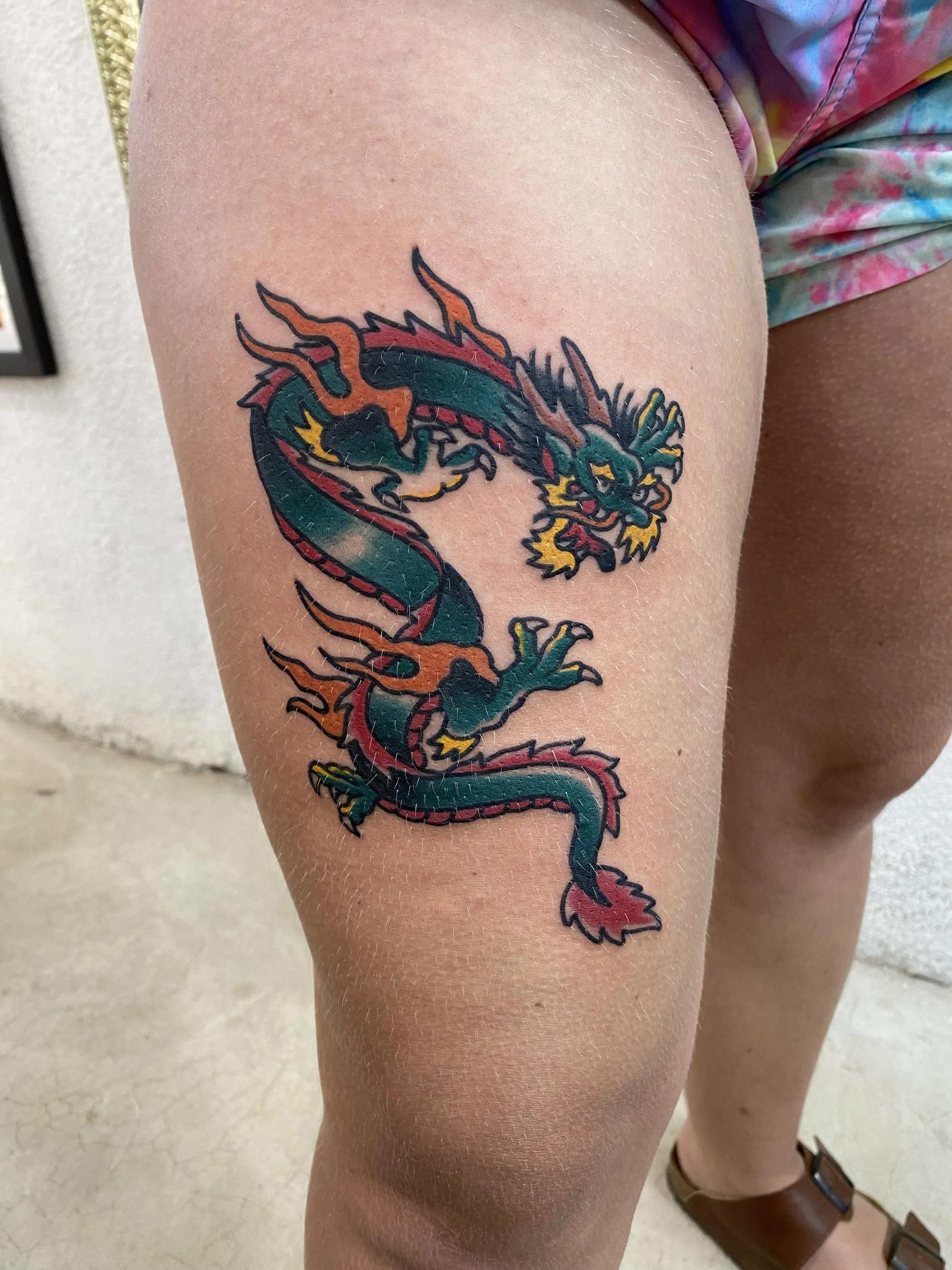 A person's thigh with a colorful tattoo of a dragon in black, red, orange, green, and yellow ink.