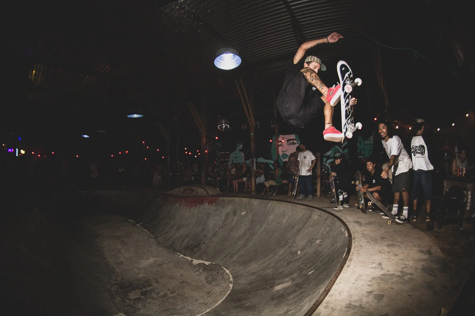 Skateboarder in mid-air performing tricks at an indoor skate park with spectators watching.