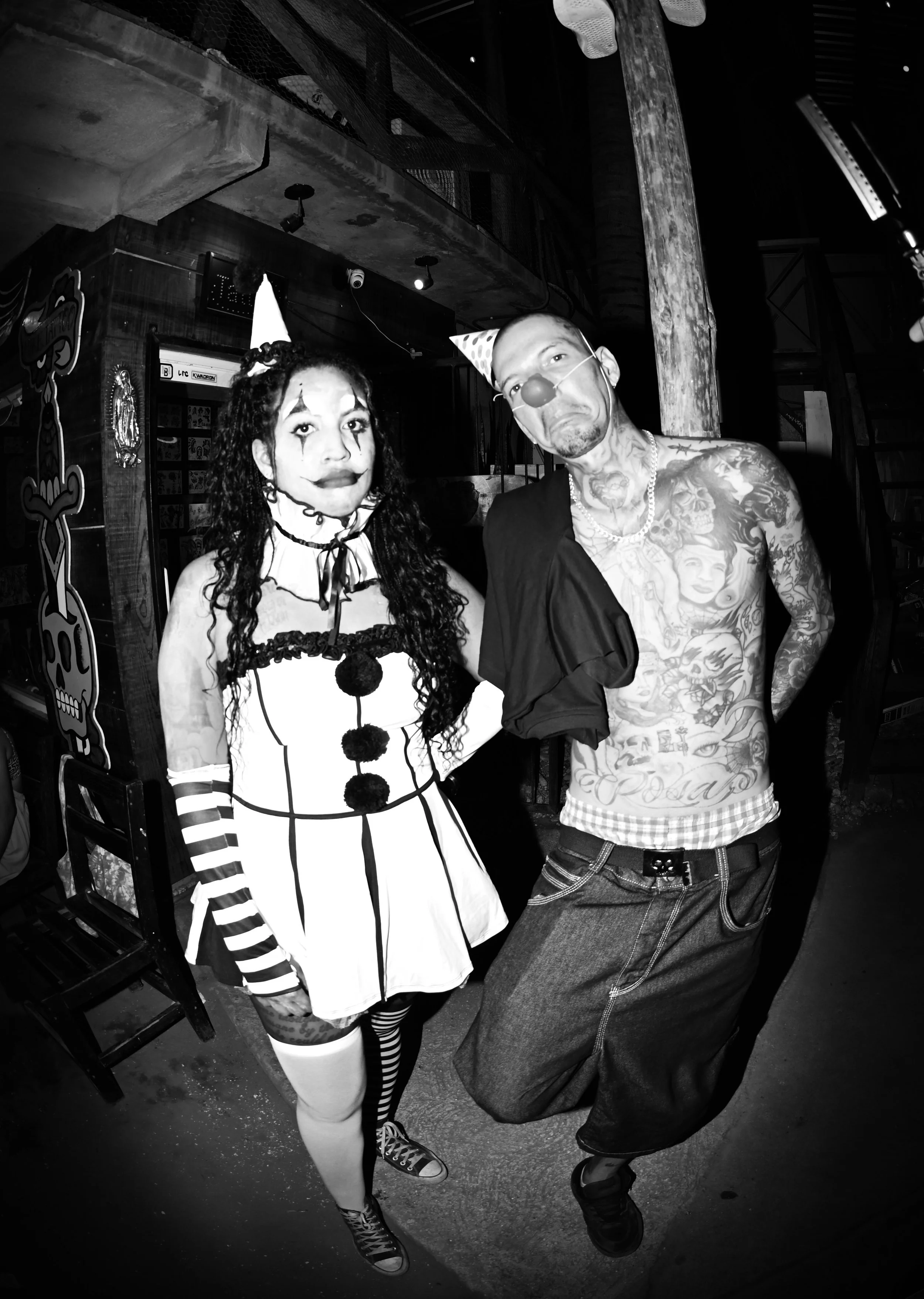 Two people dressed as clowns at a party, with one wearing a costume with black and white stripes and a hat, and the other with tattoos, a clown nose, and a party hat, both making funny faces.