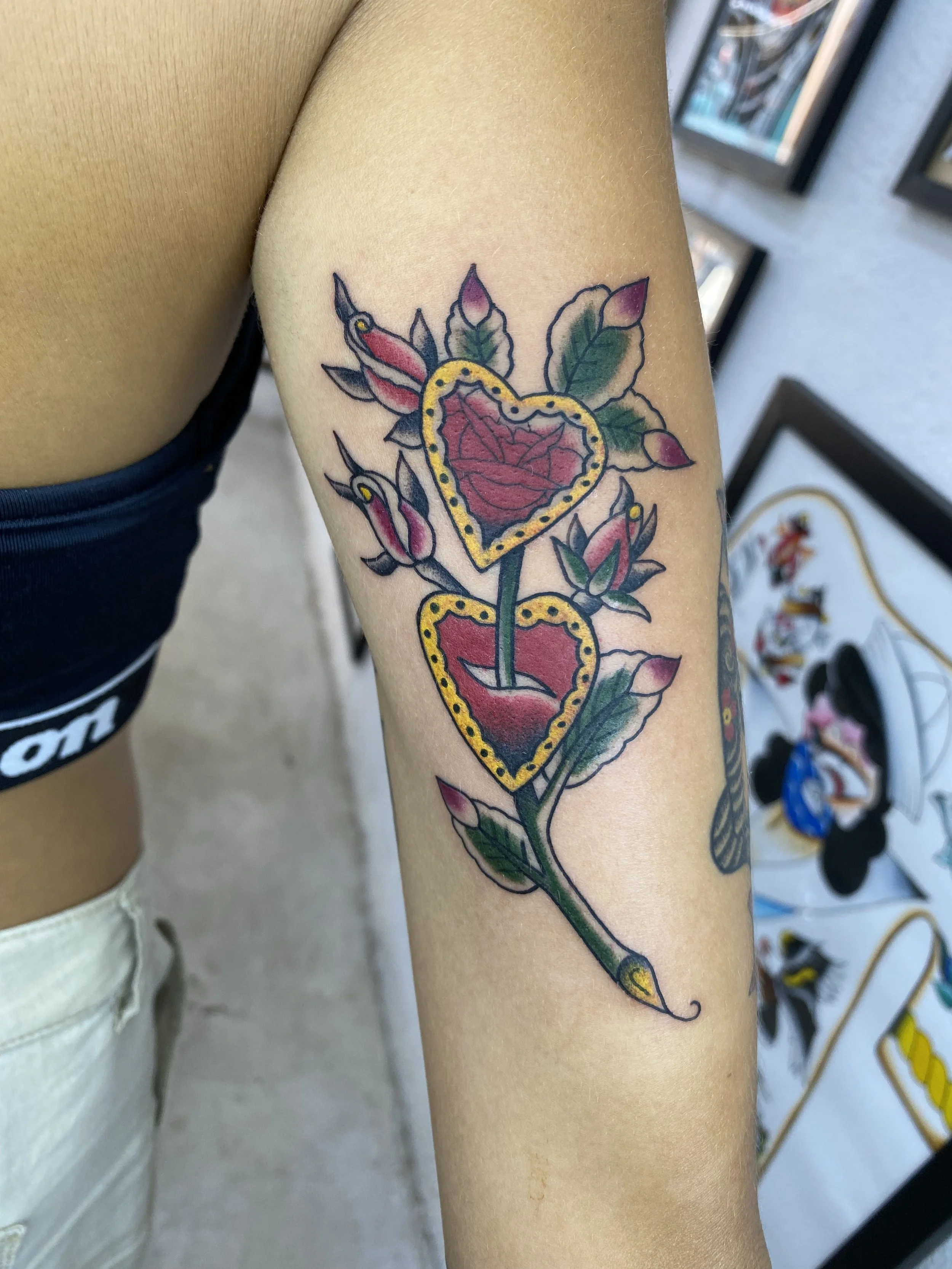 A colorful tattoo of a heart-shaped lollipop with a pink rose inside, surrounded by green leaves and pink flower buds on the person's upper arm.