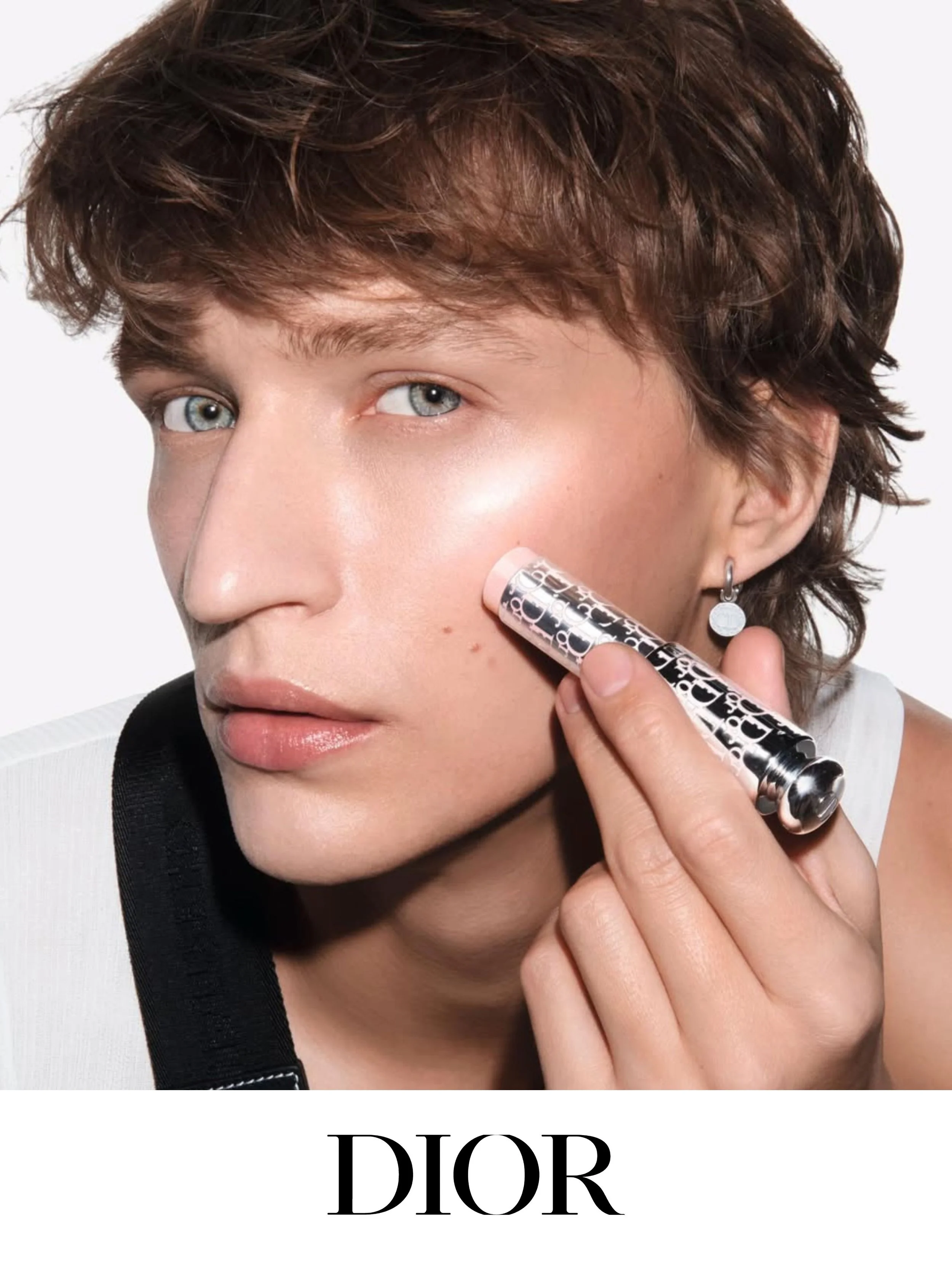 Close-up of a woman with short, curly brown hair. She is holding a silver Dior lipstick case near her cheek, with the Dior logo visible. The background is plain white, and the lower part of the image features the word 'DIOR' in large black letters.